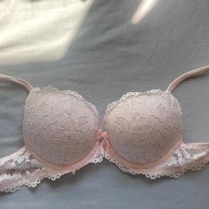 Ljusrosa spets-bh med rosett 70A, aldrig använd - Supersöt ljusrosa bh med spetsdetaljer och liten rosett framtill. Kuporna är vadderade och har blommigt spetsmönster samt dekorativ kant. Axelbanden är justerbara och bh:n har hakknäppning bak. Materialet är polyamid, elastan och polyester.