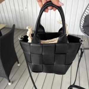 Svart flätad handväska  - Snygg svart handväska i flätad design med både handtag och justerbar axelrem. Väskan har en beige innerpåse med snörning som stängning.