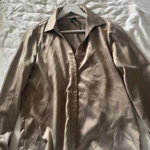 Beige skjorta i satin från Vero Moda - Stilren beige skjorta från Vero Moda i glansigt satinliknande material. Skjortan har klassisk krage, långa ärmar och knäppning framtill med matchande knappar. Säljer pga inte min storlek💖