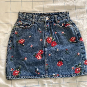 Blommig jeanskjol från Bershka - Supersöt kort jeanskjol från Bershka med broderade röda blommor över hela kjolen. Perfekt för dig som vill ha en cool och somrig vibe i garderoben!