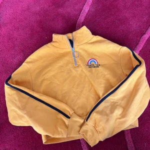 Gul half zip tröja från HM XS - Gul långärmad tröja från Divided med svart rand längs ärmarna och broderad regnbåge samt texten 'Chase the Rainbow' på bröstet. Tröjan har en snygg half zip med rund dragkedja och är gjord i mjuk bomull. Perfekt för dig som gillar färg och coola detaljer.