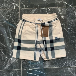 Burberry rutiga shorts med snörning - Snygga Burberry shorts i klassiskt rutigt mönster i beige, blått och svart. De har elastisk midja med vit snörning och Burberry-logga tryckt på ena benet. Perfekta för sommaren med sin lätta känsla och stilrena design.