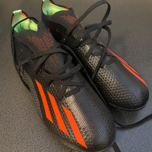 Svarta Adidas fotbollsskor - Snygga svarta Adidas fotbollsskor med röda ränder och detaljer. Ovandel i syntetmaterial med textur för bättre bollkontroll, snörning och låg profil. Insidan är ljusgrön och yttersulan har fasta dobbar för spel på gräs. 