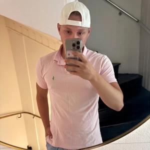 Ljusrosa pikétröja från Ralph Lauren - Säljer en ljusrosa pikétröja från Polo Ralph Lauren i custom fit. Tröjan har klassisk krage, knappar vid halsen och den ikoniska gröna loggan broderad på bröstet. Perfekt för en clean och fräsch look i sommar. Skriv innan köp så löser jag gratis frakt