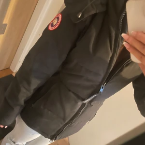 Svart kort dunjacka från Canada Goose - Säljer en svart dunjacka från Canada Goose med ikonisk patch på ärmen och stor huva med äkta päls. Jackan har dragkedja framtill, stora fickor och snygga detaljer vid huvan. Perfekt för kalla vinterdagar och riktigt varm.