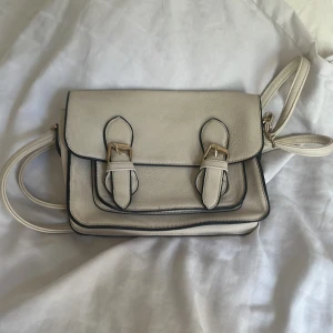 Beige axelväska med spännen - Snygg beige axelväska med två dekorativa spännen i guld och svarta kanter. Väskan har ett klassiskt, stilrent utseende och justerbar axelrem. Perfekt för att bära dina viktigaste grejer och lyfta din outfit.