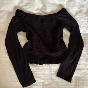 Svart smockad topp från Gina Tricot - Svart långärmad topp från Gina Tricot med smockad kropp och raka ärmar. Toppen har en lätt croppad passform och bred halsringning som kan bäras offshoulder.