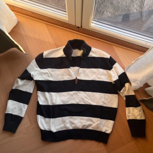 Randig stickad tröja Brandy Melville - Snygg stickad tröja från Brandy Melville med breda marinblå och vita ränder. Tröjan har hög krage, half zip-dragkedja med läderdetalj och långa ärmar. Perfekt för en chill och stilren look.