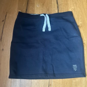 Marine Classic mörkblå kortkjol M - Mörkblå kortkjol från Marine Classic. Kjolen har en bred resår i midjan och vit snörning framtill. Tillverkad i mjuk bomull, perfekt för en avslappnad stil. Liten logga broderad nertill på framsidan.