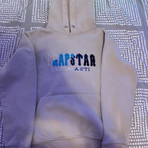 Beige Trapstar hoodie med brodyr - Säljer en beige hoodie från Trapstar med broderad logga i vitt, blått och svart framtill. Hoodien har huva med snörhål och en stor magficka. Perfekt för dig som gillar streetwear och vill sticka ut med snygga detaljer.