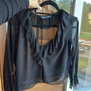 Svart volangblus från Zara XS - Säljer en svart transparent blus från Zara i storlek XS. Blusen har långa ärmar, v-ringning med volangdetaljer och är tillverkad i ett lätt, plisserat syntetmaterial. Perfekt för dig som gillar en chic och trendig look med extra detaljer.