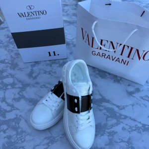 Valentino open  - Snygga vita Valentino Garavani sneakers med svart band och nitar över vristen. Skorna har rund tå, platt sula och är tillverkade i skinn. Guldiga logodetaljer på plösen och insidan. Perfekta för dig som gillar exklusiv streetstyle. Priset kan diskuteras allt ingår 