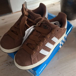 Adidas Campus 00s bruna sneakers - Adidas Campus 00s sneakers i brun mocka med klassiska vita ränder på sidorna och vit häl. Skorna har breda bruna snören och en platt, vit sula. Perfekta för dig som gillar retrovibbar och streetstyle. Modellnummer GY6433. Vit skosnöre med följs. Helt nya!!! Ord pris 1 449:- 