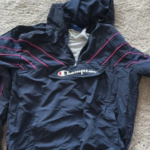 Blå vindjacka från Champion  - Snygg marinblå vindjacka från Champion med huva och halv dragkedja. Jackan har rosa detaljer och broderad logga på bröstet. Tillverkad i lätt polyester med meshfoder för extra komfort. Perfekt för en sportig och avslappnad stil.