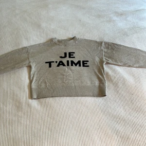 Beige tröja med text från Gina Tricot - Beige långärmad tröja från Gina Tricot med svart text 'JE T'AIME' framtill. Tröjan har rund halsringning och är i mjukt material, perfekt för en chill och stilren look.