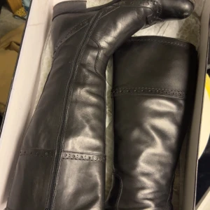 Svarta höga skinnstövlar - Snygga svarta stövlar vagabond med dekorativa sömmar och perforerade detaljer upptill. Modellen är knähög med låg klack och dragkedja på insidan för enkel på- och avtagning. Perfekta för att lyfta vilken outfit som helst.  Nypris cirka 2000kr. Dom är i fint skick