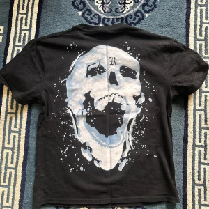 Svart Revenge t-shirt med tryck - Svart t-shirt från Revenge med stort vitt tryck på bröstet och en stor dödskalle på ryggen. T-shirten har korta ärmar och en klassisk passform. Materialet är mjuk bomull och trycket har en cool, splashad effekt som ger streetkänsla.