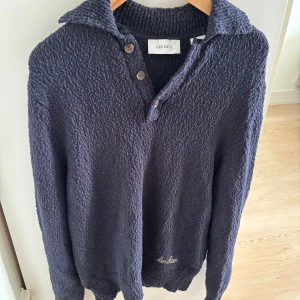 Mörkblå Les Deux tröja - Mysig mörkblå stickad tröja från Les Deux med grov struktur, krage och knappar vid halsen. Broderad logga på ärmen och ribbade muddar. Perfekt för lager-på-lager och chill dagar.