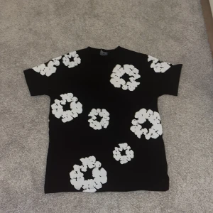 Svart blommig t-shirt från Denim Tears - Svart t-shirt från Denim Tears med stora vita blommönster över hela plagget. Klassisk passform med korta ärmar och rund hals. Perfekt statement-piece för dig som vill sticka ut med en unik och trendig look.