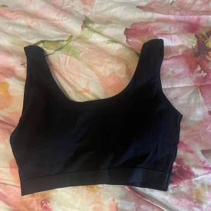 Svart ribbad linnetopp - En svart ribbad linnetopp med breda axelband och rundad halsringning. Toppen är croppad och har en enkel, stilren design som passar perfekt till jeans eller kjol. Materialet är stretchigt och bekvämt.