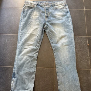 Trapstar x Adwoa jeans ljusblå. Mått står i beskrivningen👇 - Säljer ett par ljusblå jeans köpta i London från Trapstar med snygg broderad detalj på bakfickan och råa, fransiga benslut. Jeansen har dragkedjor vid bensluten med T-formad zip och klassisk femficksdesign. Perfekt för dig som gillar streetwear och unika detaljer. Midjemått: 50cm.  Innerbenslängden: 80cm. Byxlängd: 108cm.