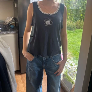 Mörkblå linnetopp med spetskant - Snygg mörkblå linnetopp med vit spetskant runt halsringningen och ett broderat blommotiv framtill. Avslappnad passform och ärmlös design, perfekt för en chill look. Passar till både jeans och kjol.