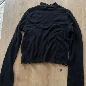 Svart finstickad polotröja med volangkant - Svart långärmad polotröja med volangkant vid hals, ärmslut och nederkant. Tröjan är tight och har en mjuk, stretchig känsla som passar perfekt till jeans eller kjol. Enkel men ändå med en söt detalj tack vare volangerna.