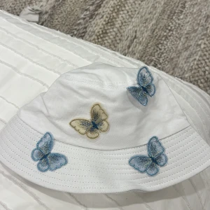 Vit bucket hat med fjärilar - Superfin vit bucket hat dekorerad med broderade fjärilar i blått och beige. 