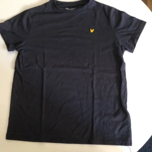 Mörkblå t-shirt från Lyle & Scott - Snygg mörkblå t-shirt från Lyle & Scott med klassisk passform och korta ärmar. T-shirten har en gul broderad logga på bröstet och är gjord i mjuk bomull. Perfekt för en clean och stilren look.