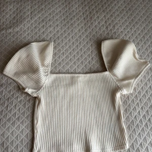 Ribbad offshoulder topp från H&M - Superfin ribbad offshoulder topp från H&M i ljusbeige. Toppen har korta, vida ärmar och croppad passform som ger en trendig look. Perfekt att styla med höga jeans eller kjol för en cool vibe.