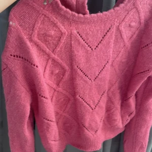 Rosa stickad tröja med mönster - Mysig rosa stickad tröja med snyggt hålmönster och geometriska former på framsidan. Tröjan har v-ringning med två knappar och långa ärmar. Perfekt för dig som gillar färg och vill ha en unik detalj i garderoben.