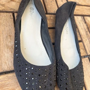Svarta ballerinaskor med nitar från Znyshoes - Svarta ballerinaskor från Znyshoes med snygga svarta nitar på ovandelen. Skorna har rund tå, platt sula med blommönster undertill och är gjorda i mockaliknande material. Perfekta för dig som vill ha en stilren men ändå lite edgy look.