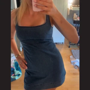 Mörkblå jeansklänning med smala axelband - Säljer en mörkblå jeansklänning med smala axelband och fyrkantig urringning. Klänningen är figurnära och har sömdetaljer under bysten för en snygg passform. Perfekt för dig som gillar en enkel men trendig look.