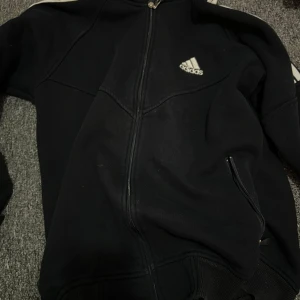 Svart Adidas jacka med dragkedja - Svart Adidas jacka med klassiska vita ränder längs ärmarna och stort Adidas-tryck på ena ärmen. Jackan har hel dragkedja framtill, ribbade muddar och två fickor med dragkedja. Perfekt för en sportig look och chill vardagsstil.