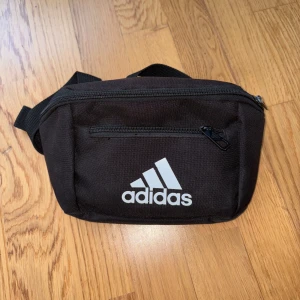 Svart magväska från Adidas - Säljer en svart magväska från Adidas med klassisk logga framtill. Väskan har justerbart band, dragkedja upptill och ett extra fack med dragkedja på framsidan. Perfekt för att bära mobilen, plånboken och andra smågrejer när du är på språng.
