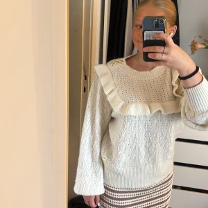 Vit stickad tröja med volang Holly & White - Supersöt vit stickad tröja från Holly & White med volangdetalj över bröstet och axlarna, samt guldknappar på ena axeln. Tröjan har ett hålmönster upptill och bred ribbad mudd nertill och vid ärmslut. Perfekt för dig som gillar romantiska detaljer.