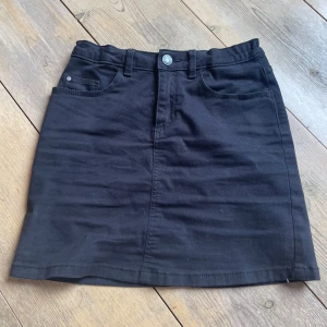 Svart jeanskjol - Svart jeanskjol från Cubus med klassiska fickor, dragkedja framtill och resårband i midjan. Storlek 146/152 men skulle även argumentera för att det passar en xxs-xs!!