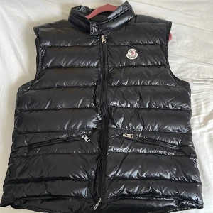 Svart dunväst från Moncler - Snygg svart dunväst från Moncler med glansig finish och klassisk logga på bröstet. Västen har hög krage, två dragkedjefickor och kraftig dragkedja framtill. Perfekt för lager-på-lager och riktigt street. Fodrad med dun för extra värme.