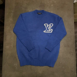 Blå stickad tröja Louis Vuitton - Snygg blå stickad tröja från Louis Vuitton med stort vitt LV-logo på bröstet. Tröjan har rund hals, ribbade muddar och är långärmad. Perfekt för dig som vill ha en exklusiv och trendig look!