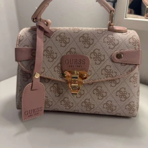 Beige handväska från Guess med monogram - Snygg beige handväska med klassiskt monogrammönster och detaljer i ljusrosa. Band tillkommer.💕