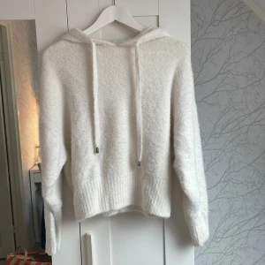Vit fluffig hoodie från H&M - Supermjuk och fluffig vit hoodie från H&M i storlek S. Tröjan har huva med snörning och långa ärmar. Den är stickad och har en mysig känsla, perfekt för kyliga dagar. Ribbstickad nederkant och ärmslut ger en snygg finish.
