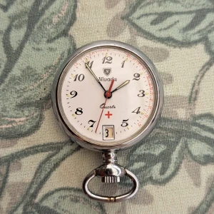 Silver fickur från Nivada Quartz - Klassiskt runt fickur från Nivada med vit urtavla, svarta siffror och röda detaljer. Urtavlan har datumfönster och röd sekundvisare. Boetten är i silverfärgat metall och har en tydlig ögla för kedja. Perfekt för dig som gillar vintage och stilrena accessoarer.