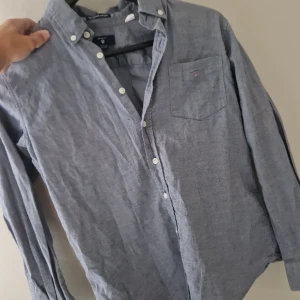 Blå oxfordskjorta från Gant - Klassisk blå oxfordskjorta från Gant med button down-krage och vita knappar. Bröstficka med broderad Gant-logga och sköld. Skjortan är långärmad och i mjuk bomull, perfekt för en clean och stilren look.