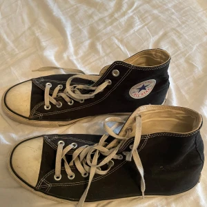 Svarta Converse Chuck Taylor All Star - Klassiska svarta Converse Chuck Taylor All Star med högt skaft. Skorna har vit gummitå, vita snören och tydliga kontrastsömmar. Ikonisk logga på sidan och slitstark gummisula. Perfekta för en avslappnad och cool streetstil. ACCEPTERAR TYP ALLA PRISFÖRSLAG 
