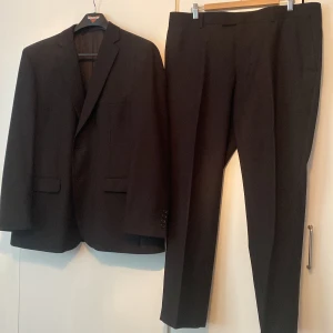Svart kostym med kavaj och byxor - Klassisk svart kostym från Dressman med stilren kavaj och matchande kostymbyxor. Kavajen har två knappar, fickor med lock och smal krage. Byxorna är raka med pressveck och hällor för bälte. Perfekt för dig som vill ha en tidlös och snygg look.                                 Kavajen- stl 56 short.  Byxorna- W38 L 30