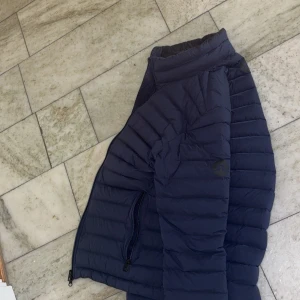 Mörkblå dunjacka från J.Lindeberg - Snygg mörkblå dunjacka från J.Lindeberg med horisontella sömmar och diskret logga på ärmen. Jackan har dragkedja framtill och praktiska fickor med dragkedja. Perfekt för kyliga dagar och enkel att matcha med olika outfits.