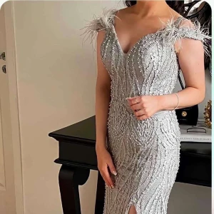 Silverglittrig långklänning med fjädrar - Magisk långklänning i silver med glittriga paljetter och pärlor, v-ringad och med fjäderdetaljer på axlarna. Klänningen har en snygg slits framtill och figurnära passform, perfekt för bal eller fest. Materialet är glansigt och skimrande, vilket ger en lyxig känsla. Den är använd en gång!