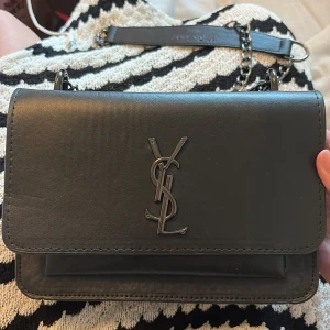 Svart axelväska från Saint Laurent - Snygg svart axelväska från Saint Laurent med klassisk YSL-logga i metall framtill. Väskan har kedjerem i metall och är tillverkad i slätt skinn. Perfekt storlek för det viktigaste och ger en lyxig touch till din outfit. 