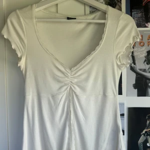 Vit omlottopp med snörning Gina Tricot - Supersöt vit topp från Gina Tricot med v-ringning, snörning framtill och fina spetsdetaljer längs kanterna. Kortärmad modell med lätt rynk vid bysten och mjukt, stretchigt material. Perfekt för dig som gillar romantisk och feminin stil.