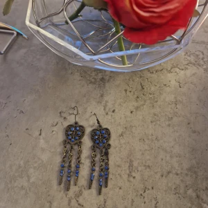 Hjärtformade örhängen med blå detaljer - Unika örhängen i antikguld med hjärtform och blå stenar. Hängande kedjor med blå pärlor och spetsiga detaljer ger en cool boho-vibe. Perfekta för dig som vill sticka ut med accessoarer som har både färg och attityd.